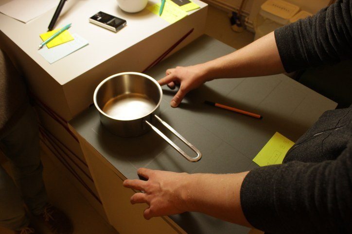 Hands placing a small pan on a cardboard mock-up of a cooktop, with paper sketches next to it
