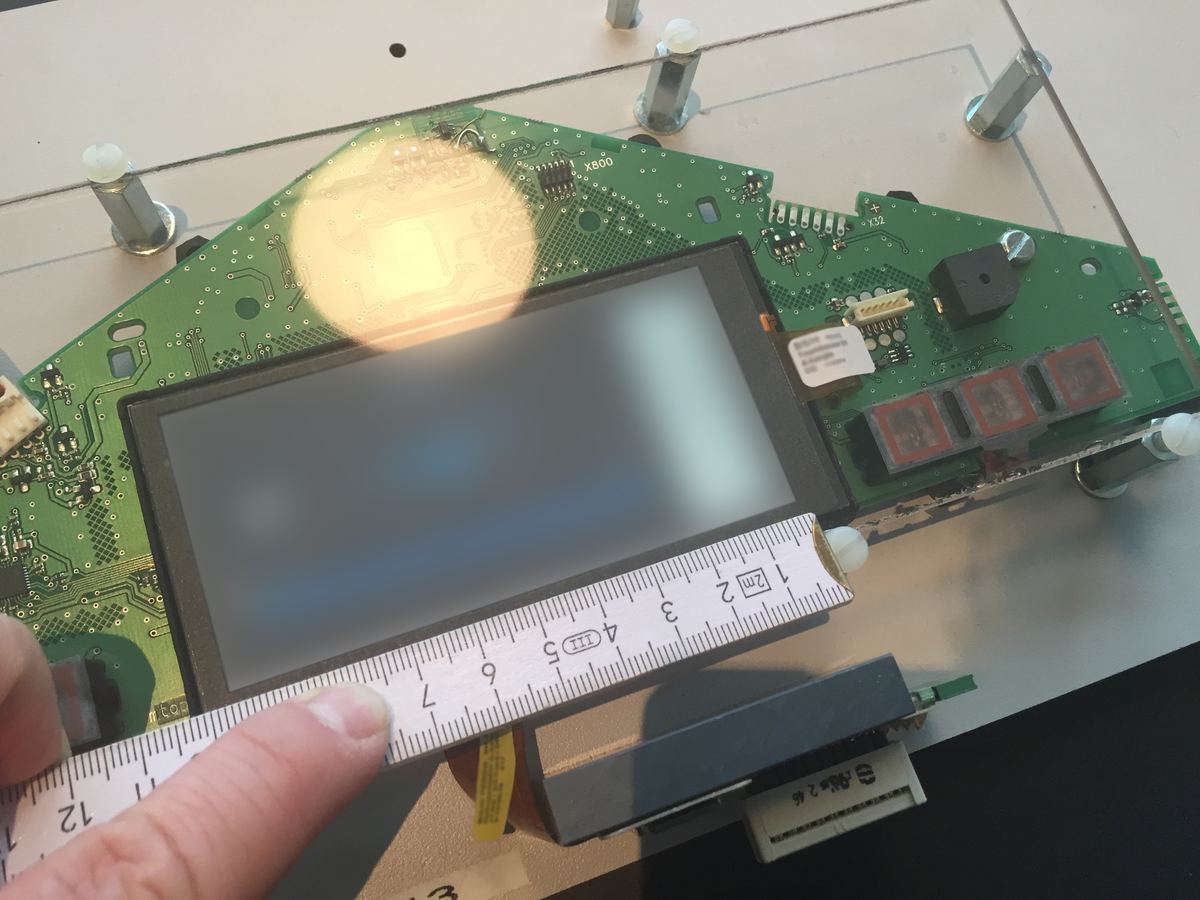 A hand holding a ruler against a display on a green circuit board; the display content is blurred