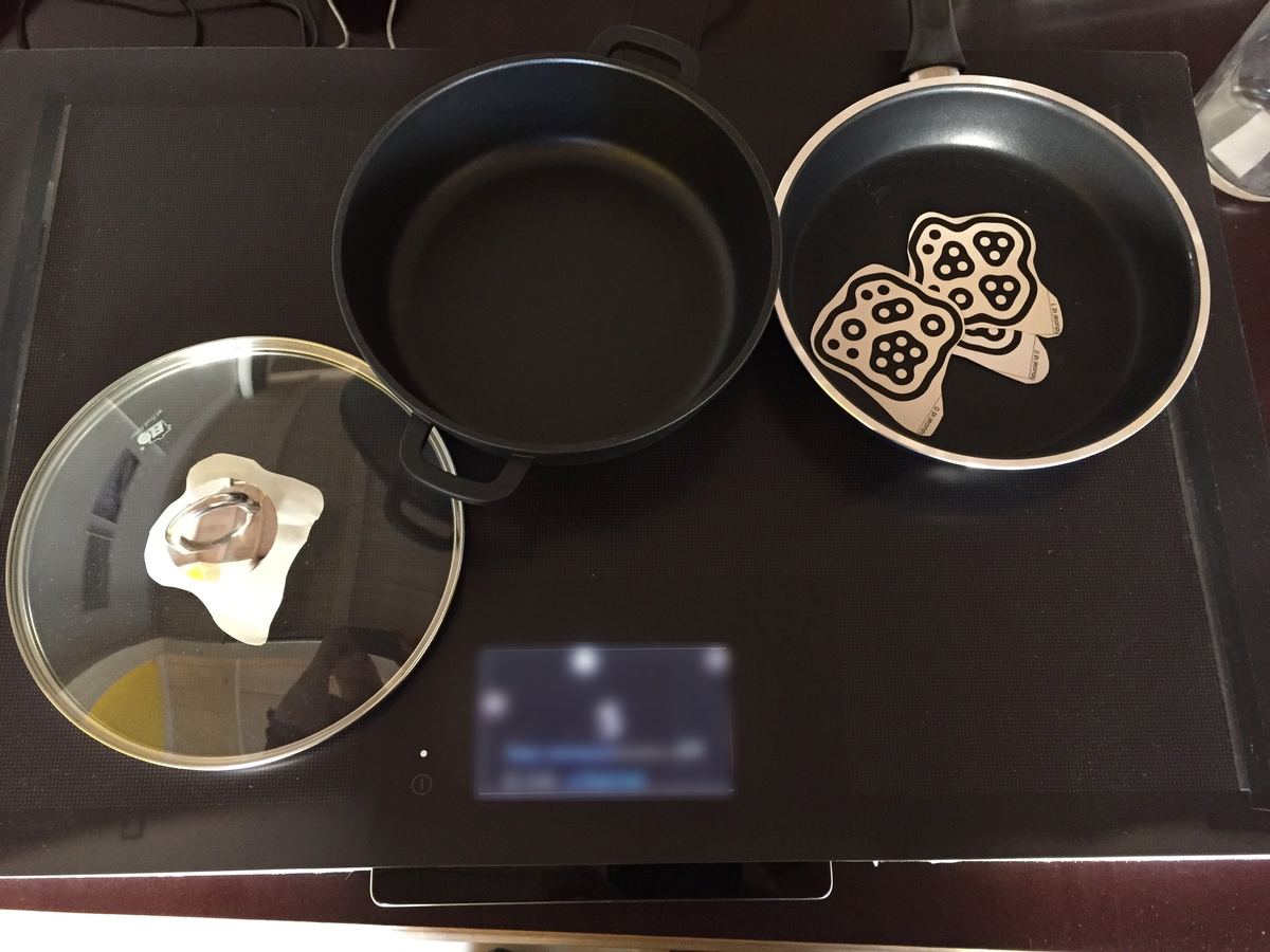 Pans and a lid with markers on a dark prototype cooktop; the small display below is blurred